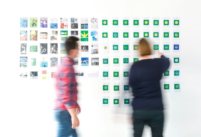 Dwie osoby przed ścianą z wydrukami projektów i wariantami identyfikacji wizualnej.